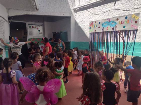 Animador fantasiado com chapéu colorido dança com as crianças fantasiadas, em frente ao painel “Festa da Alegria”