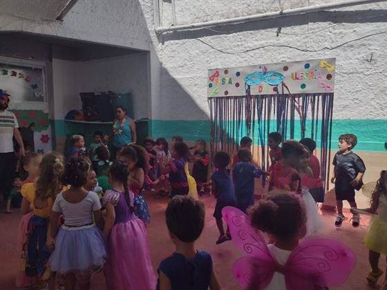 Crianças fantasiadas dançam no pátio, com o painel “Festa da Alegria” e máscaras de carnaval ao fundo