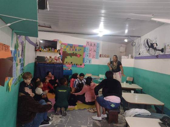 Crianças e familiares sentados no chão da sala assistindo a uma contação de histórias com teatrinho