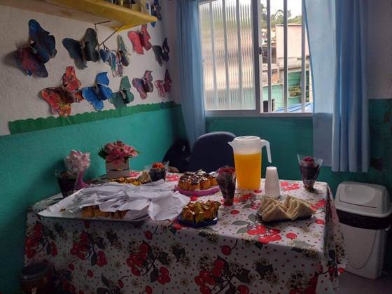 Mesa de lanche com bolos, salgados e suco, ao lado de uma parede decorada com borboletas feitas pelas crianças