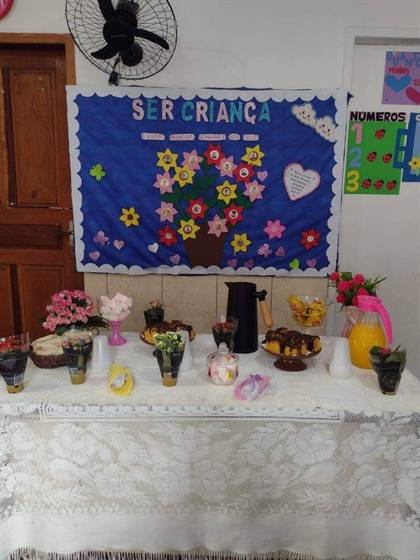 Mesa de lanche com bolos, docinhos e suco, em frente ao mural “Ser Criança é viver, brincar, sonhar e ser feliz”