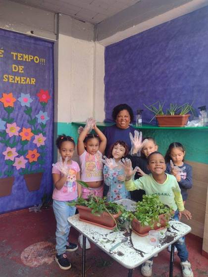 Crianças de luvas plásticas mostrando as mãos ao lado de uma educadora, em frente a jardineiras com mudas