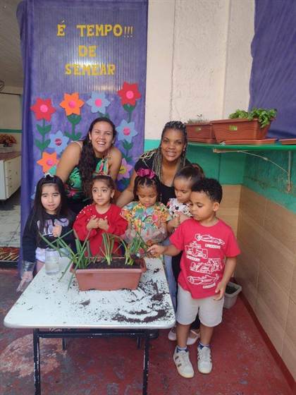 Duas educadoras e cinco crianças reunidas em volta de uma jardineira com mudas, no painel “É tempo de semear”