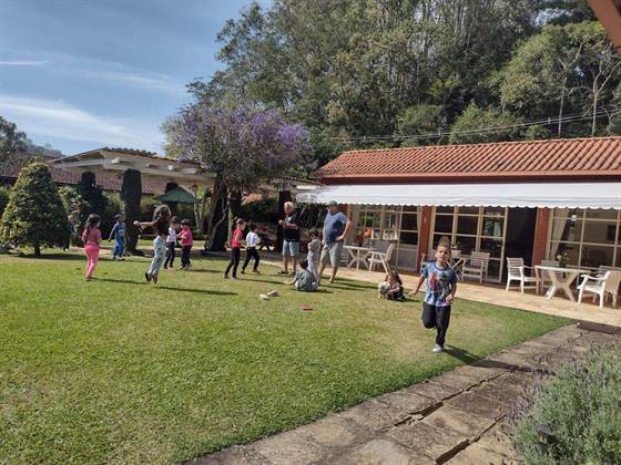 Crianças correndo no gramado em frente à casa, com um pé de quaresmeira florido e um cachorrinho por perto