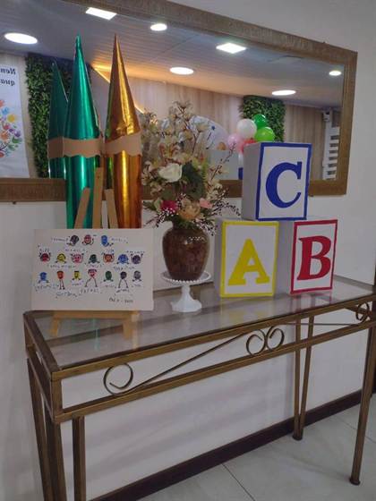Mesa de recepção com lápis gigantes, cubos ABC, flores e quadro de digitais da turma Pré 2 de 2025
