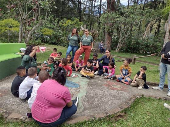 Crianças sentadas em roda ao ar livre durante uma atividade educativa com as guias do Horto