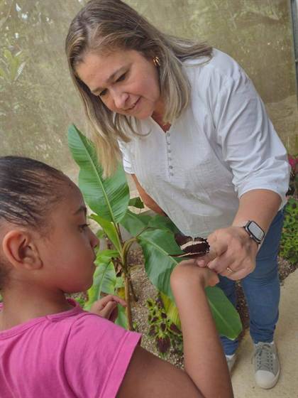 Educadora segura uma borboleta no braço de uma criança, dentro do Borboletário