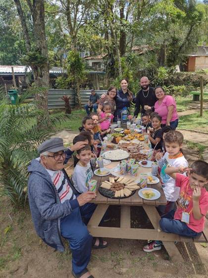 Outro ângulo da mesa de piquenique com lanche, crianças e acompanhantes, no Horto