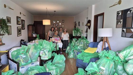Voluntárias, as ajudantes do Papai Noel, sorrindo atrás de uma mesa, cercadas por sacolas verdes com presentes e brinquedos