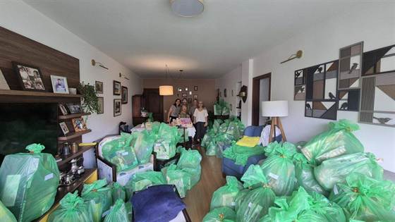 Sala tomada por dezenas de sacolas verdes com presentes e brinquedos, com cinco pessoas ao fundo